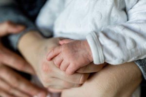 Adult arms embracing small baby. In focus are a small baby hand on top of an adult hand.