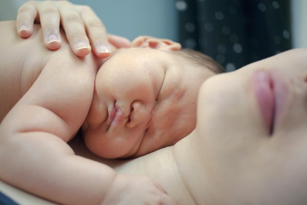 Naked baby lies asleep on their mother's chest. Mother has an arm wrapped around her baby.