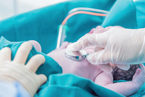 A healthcare professional uses a stethoscope to examine a newborn shortly after birth in a clinical setting. The infant is lying on a teal surgical drape, and the scene appears to be within a neonatal intensive care unit. The newborn is receiving immediate medical attention, with gloved hands gently conducting the examination.