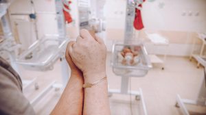 Two people holding hands in front of a neonatal incubator with a newborn baby inside, in a hospital NICU.