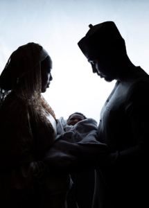 Silhouetted African couple lovingly holding a newborn baby wrapped in a blanket, standing face-to-face against a soft backlight.